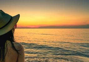 vista traseira da mulher sexy assistindo o pôr do sol na praia. mulher usa óculos escuros e chapéu de palha relaxante na praia do paraíso tropical. férias de verão. viagens de férias sozinho. Ritmo de verão. lindo céu por do sol