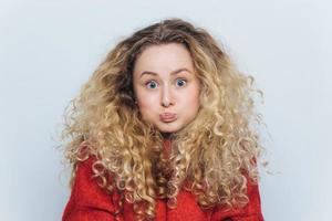 mulher loira fuuny com cabelo encaracolado, sopra as bochechas e olha para a câmera, reage ativamente em notícias inesperadas, isoladas sobre fundo branco. jovem emocional posa no interior. conceito de linguagem corporal foto