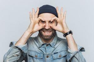 retrato de homem barbudo hppy faz óculos com os dedos, tem barba por fazer, usa jeans coagulação, fica contra um fundo branco. engraçado homem envelhecido de meia-idade interior, finge usar óculos. foto