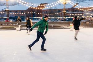 alegre homem barbudo pratica patinar no ringue de gelo, tem expressão alegre, sorri alegremente, demonstra seu profissionalismo. homem esportivo ativo na jaqueta verde vai para o esporte de inverno foto