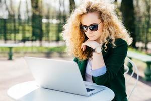 jovem mulher com cabelo loiro encaracolado usando óculos escuros e elegante jaqueta verde sentado na frente do laptop aberto ao ar livre lendo notícias on-line com expressão pensativa. pessoas, conceito de tecnologia