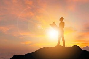 conceito de felicidade de sucesso de celebração, silhueta de homem de engenheiro de negócios segurando impressão azul pensar e olhando para a frente em um fundo de céu de noite de pôr do sol no topo da montanha com traçado de recorte. foto