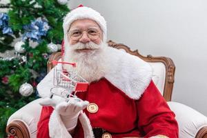 papai noel fazendo compras no supermercado, ele está mostrando um mini carrinho, natal e conceito de compras. foto