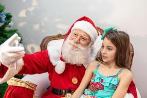 papai noel sentado com uma garota no colo fazendo fotos de selfie com seu celular.