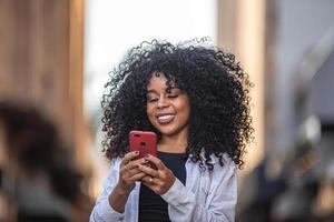 jovem negra de cabelo encaracolado andando usando o celular. mensagens de texto na rua. cidade grande. foto