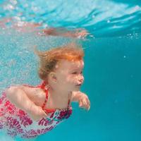 menina sorridente no vestido moderno bonito mergulho debaixo d'água na piscina azul. estilo de vida ativo, aula de natação infantil com os pais. atividade de esportes aquáticos durante as férias de verão em família no resort foto