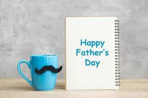 feliz dia dos pais com o bloco de notas de papel, xícara de café azul ou caneca de chá e decoração de bigode preto na mesa. dia internacional dos homens e conceito de celebração foto