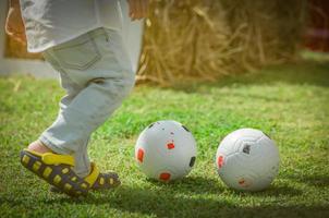 menino bonito feliz jogando futebol fora de casa ou escola em dia de verão. criança pré-escolar joga futebol no gramado da grama verde. criança com sapatos casuais pratica esporte no parque. um menino divertido com jogo de futebol sozinho foto