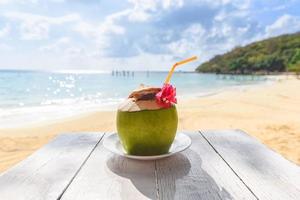 frutas tropicais de coco na mesa e areia fundo de praia água - verão de suco de coco fresco com flor no mar da praia em clima quente paisagem do oceano natureza férias ao ar livre, coco jovem foto