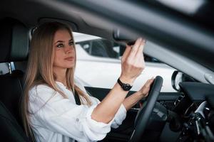 usa espelho para dar uma olhada em si mesma. linda loira sentada no carro novo com interior preto moderno foto