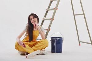 o que pintar a seguir. linda morena de uniforme amarelo e balde de cor azul fica perto da escada foto