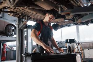 procurando os instrumentos necessários. homem na oficina de uniforme conserta peças quebradas do carro moderno foto