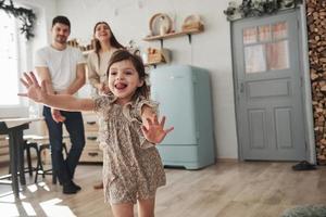 daqui para frente. criança do sexo feminino brincalhão se diverte correndo na cozinha durante o dia na frente de sua mãe e pai foto