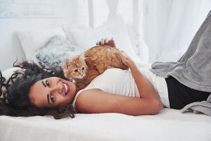 garota está sorrindo, gato está olhando para o lado. loira atraente, descansando na cama branca com seu gatinho fofo foto