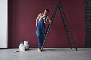 encostado na escada. jovem trabalhador afro-americano de uniforme azul tem algum trabalho a fazer foto