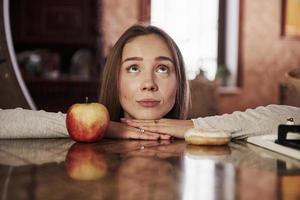 dona de casa jovem bonita inclinando-se sobre a mesa com maçã e biscoito e olha para cima foto