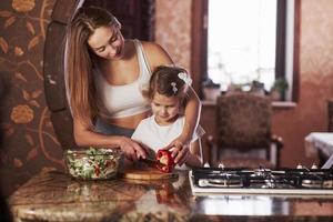 você pode facilmente fazê-lo. bela jovem de pé na cozinha moderna perto do fogão a gás e ensina a filha a preparar comida foto