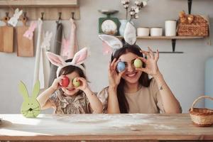 sorrindo e segurando ovos. mãe e filha em orelhas de coelho na páscoa se divertem na cozinha durante o dia