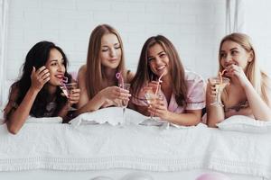 bebendo alguns bons coquetéis. meninas alegres de pijamas deitadas na cama no quarto branco e celebram foto