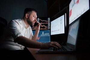 esclarece as informações. homem barbudo de camisa branca tem uma ligação comercial e trabalha no escritório com várias telas de computador em gráficos de índice foto