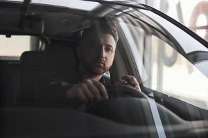 relaxando com charuto na mão. vista frontal do jovem empresário barbudo sentado em seu carro preto de luxo foto