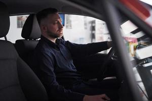 na camisa preta. vista lateral do jovem empresário barbudo sentado em seu carro vermelho de luxo foto