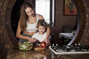 tenta cortar vegetais. bela jovem de pé na cozinha moderna perto do fogão a gás e ensinar a filha a preparar comida foto