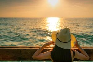 vista traseira da mulher asiática sexy de cabelo comprido preto com chapéu de palha relaxando e curtindo as férias na praia do paraíso tropical e assistir o céu pôr do sol. férias de verão. viagem de férias sozinho. Ritmo de verão. foto