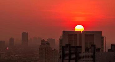 paisagem urbana de manhã com céu ao nascer do sol e poluição do ar. poeira fina de pm 2.5 cobre a cidade. paisagem urbana com prédio residencial lotado. nascer do sol urbano com grande sol vermelho. fumaça no ar. pouco saudável.