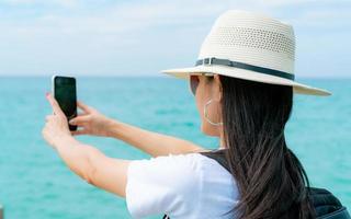 mulher jovem mochileiro asiático usar chapéu usar smartphone tomando selfie no cais. férias de verão na praia do paraíso tropical. garota hipster feliz viaja de férias. mulher aproveite e relaxe a vida. Ritmo de verão. foto