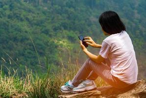 jovem mulher asiática sente-se na rocha no penhasco e usando mensagem de telefone inteligente para seus amigos durante as férias no parque nacional. conceito de viagem e comunicação de aventura foto