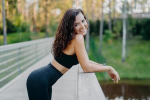 tiro lateral de mulher desportiva magro usa top preto e leggings, tem uma figura perfeita, inclina-se na ponte, faz uma pausa depois de fazer exercícios esportivos, leva um estilo de vida saudável. corredor feminino gosta de natureza foto