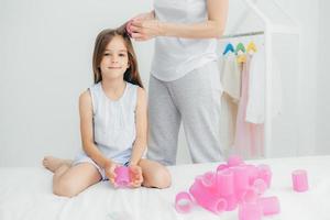 foto recortada da mãe irreconhecível enrola bobes para sua filha bonita, vai fazer um belo penteado, tem uma aparência agradável e cabelos longos. adorável adorável menina quer ter cachos