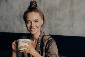 retrato de jovem morena atraente com lindo sorriso largo bebendo café na cama foto