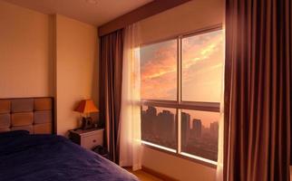 quarto de manhã com a luz do sol da manhã através da janela de vidro com cortinas abertas. cama de luxo em apartamento moderno na cidade. interior do quarto. cama coberta de cobertor azul. vista da janela de vidro. foto