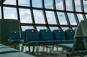 assento vazio na sala de embarque no terminal do aeroporto. distância para um assento mantenha distância para proteger o coronavírus e o distanciamento social dos passageiros por segurança. visto avião voando pela janela de vidro. foto