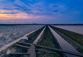 fazenda de sal de manhã com céu e nuvens ao nascer do sol. paisagem do campo de sal marinho na tailândia. água do mar no canal e caminho do solo na fazenda. matéria-prima de sal industrial. turismo de verão na tailândia. foto