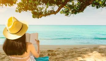 as mulheres sentam e lêem um livro debaixo da árvore à beira-mar. vista traseira da mulher asiática sexy com chapéu de palha relaxando e aproveitando as férias na praia de areia do paraíso tropical. férias de verão. Ritmo de verão. foto