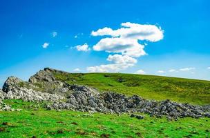 paisagem de grama verde e colina de pedra na primavera com lindo céu azul e nuvens brancas. campo ou vista rural. fundo da natureza em dia ensolarado. ambiente de ar fresco. pedra na montanha. foto