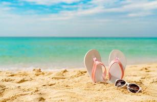 sandálias rosa e brancas, óculos de sol na praia de areia à beira-mar. chinelos e óculos de estilo de moda casual. férias de verão na praia tropical. divertidas viagens de férias na praia. horário de verão. Ritmo de verão. foto
