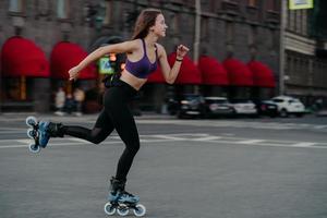 tiro horizontal de jovem magro ativo anda rápido em patins melhora a agilidade do equilíbrio e a coordenação vestida com top recortado e poses de legging na estrada da rua focada na distância foto