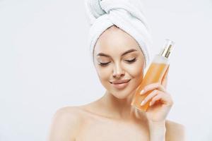 tiro horizontal de mulher bonita com pele saudável, maquiagem natural, usa spray de perfume, tem expressão satisfeita, beleza natural, olha para baixo, usa toalha de banho na cabeça, isolada sobre fundo branco foto