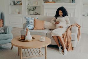 mulher afro-americana grávida sentada no sofá e passando tempo em casa foto