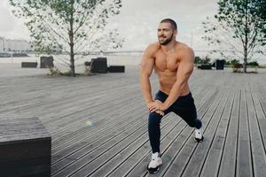 tiro horizontal de homem barbudo motivado estica o músculo da perna antes de exercícios esportivos ou corrida, aquece, posa no parque da cidade ao ar livre, tem torso musculoso. esportista pratica esportes regularmente foto