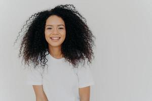 tiro na cabeça da mulher sorridente tem cabelo espesso luminoso, fica confiante e satisfeito, maquiagem mínima, gosta de uma conversa animada, vestida com uma confortável camiseta branca, posa no interior. emoções felizes. foto