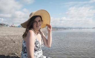 bela jovem hispânica feliz sentada sozinha na beira da praia olhando para a câmera, usando um chapéu e um vestido preto e branco durante uma manhã ensolarada foto