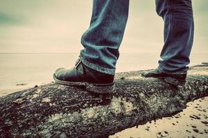 homem de jeans e sapatos elegantes em pé na árvore caída na praia selvagem, olhando para o mar. vintage foto