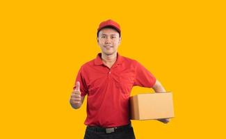 trabalhador de entregador asiático sorrindo e polegares para cima em uniforme vermelho isolado em fundo amarelo, segure caixas de encomendas para enviar ou transportar encomendas por correio. foto