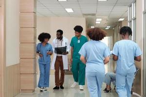grupo de praticantes, médico afro-americano profissional com equipe de jovens estudantes de medicina, caminhada feliz e discutir filme de raio-x de diagnóstico no exame ambulatorial de saúde no hospital.