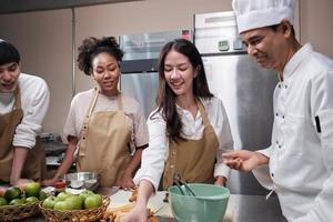 curso de culinária, chef masculino sênior em uniforme de cozinheiro ensina jovens estudantes de aula de culinária a amassar e enrolar massa de pastelaria, preparar ingredientes para alimentos de padaria, tortas de frutas na cozinha de aço inoxidável. foto